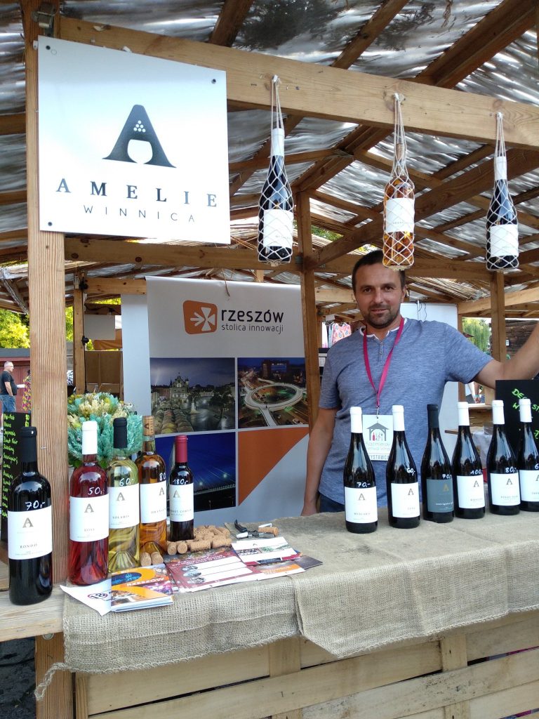 Tablica z logo Winnicy Amelie i butelki wina w pergoli — degustacje i wydarzenia